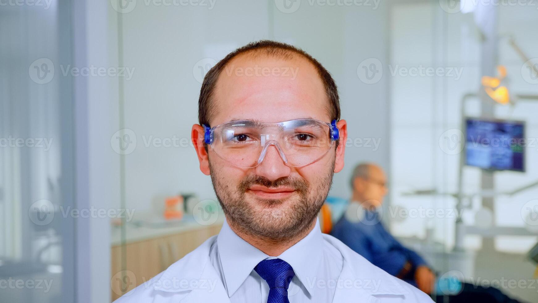 Dentist doctor looking at camera smiling while elderly patient waiting him in background for dental hygiene. Stomatologist with protection glasses standing in front of webcam in stomatological clinic. photo