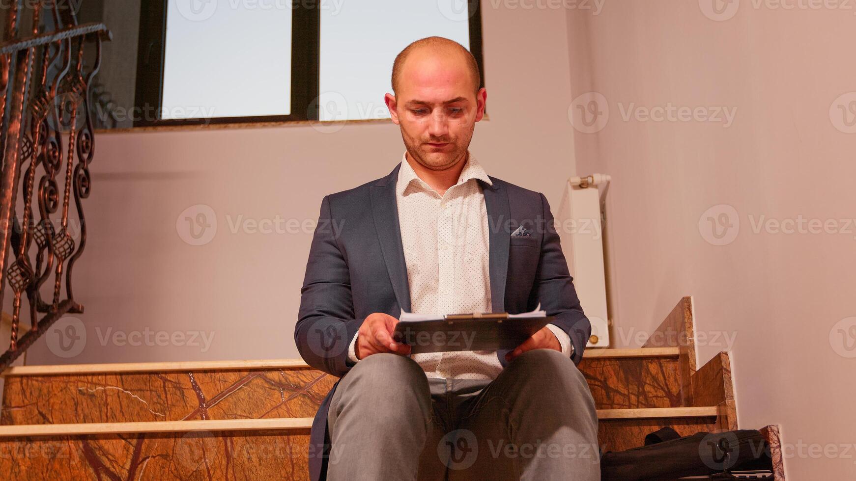 Businessman working on stairs in finance corporate company checking documents holding clipboard while colleagues moving on staircase. Professional businesspeople in modern financial building. photo