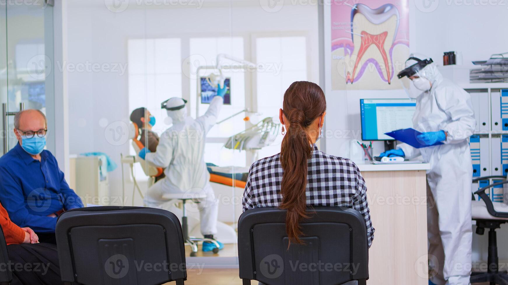 Assistant with face mask against covid-19 coming in reception typing on computer while patients sitting on chairs in waiting room talking keeping distance. Concept of new normal dentist visit. photo