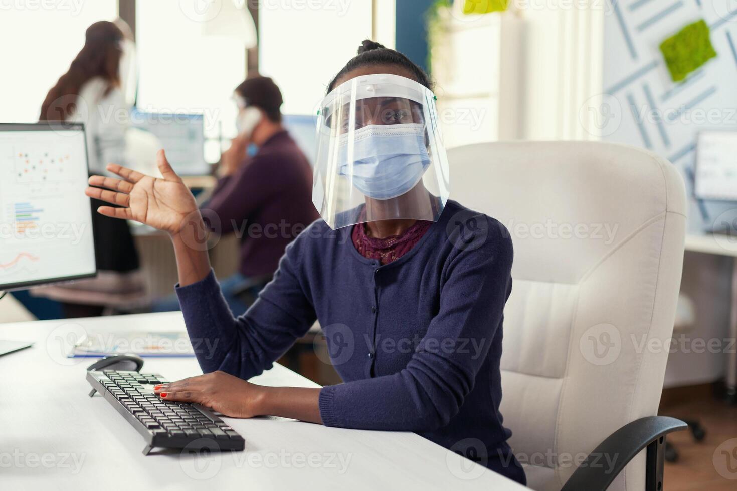 Pov of african entrepreneur waving on video call with business people weaing face mask against covid19. Woman speaking with team during online conference while colleagues working in background. photo