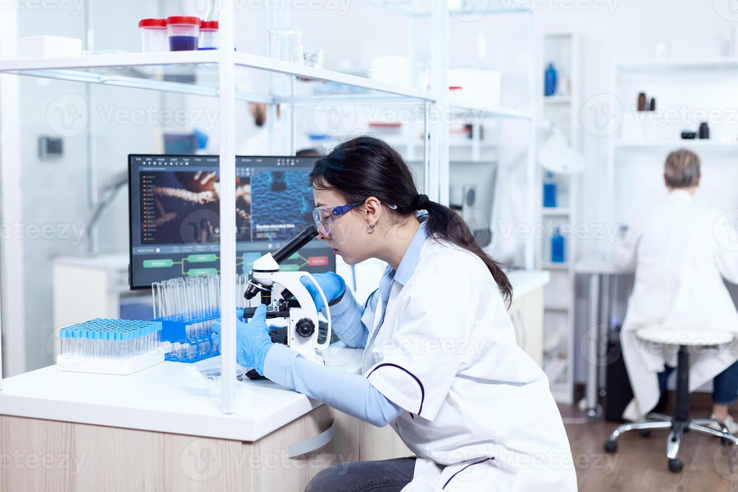 Expert in genetics doing research using microscope to analise chemical from human adn. Chemist wearing lab coat using modern technology during scientific experiment in sterile environment. photo