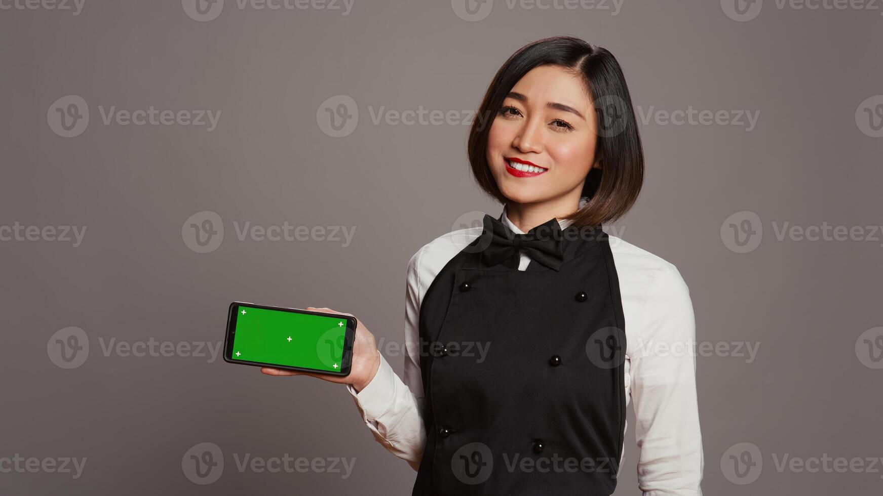 Catering employee holding phone with greenscreen display, pointing at chromakey design on smartphone. Asian waitress using isolated copyspace template over grey background in studio. Camera B. photo