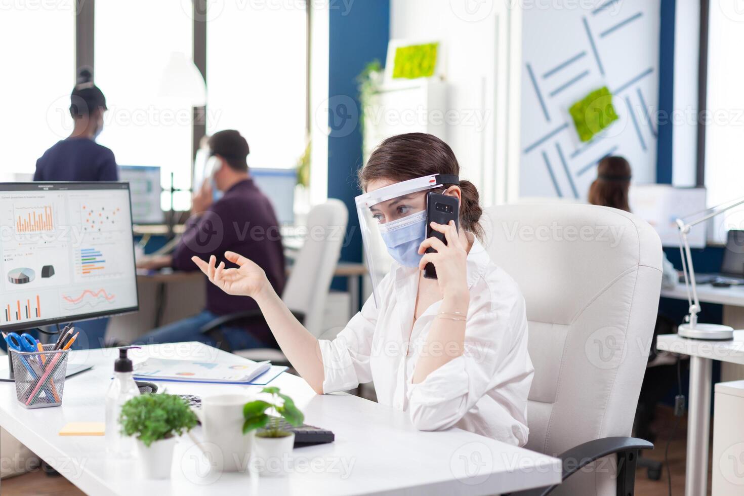 Business woman during global pandemic with covid 19 having a business conversation using smartphone wearing face mask. Multiethnic coworkers working respecting social distance in financial company. photo