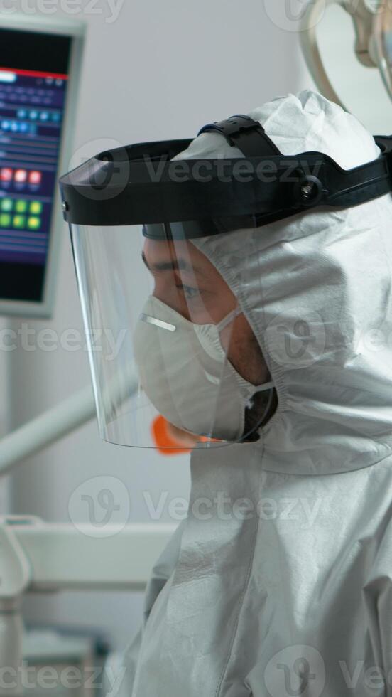 Portrait of dentist with protection mask speaking with elderly patient during global pandemic. Orthodontic doctor wearing protective suit, coverall, face shield and gloves in coronavirus period. photo