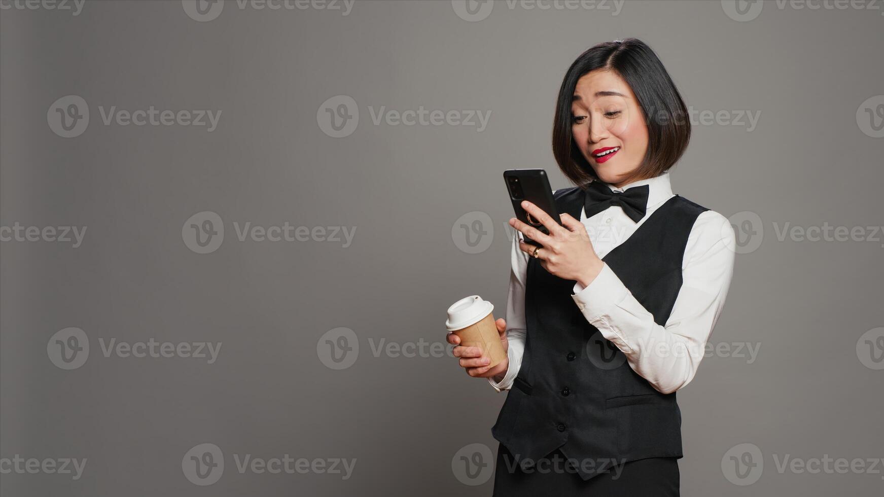 Concierge personnel drinking cup of coffee in studio, browsing smartphone social media apps. Asian receptionist enjoying caffeine refreshment while she uses mobile phone. Camera A. photo