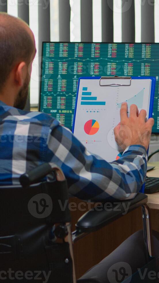 Handicapped businessman working with financial statistics checking graphs sitting immobilized in wheelchair on desk in building office. Paralysed freelacer using modern technology and documents. photo