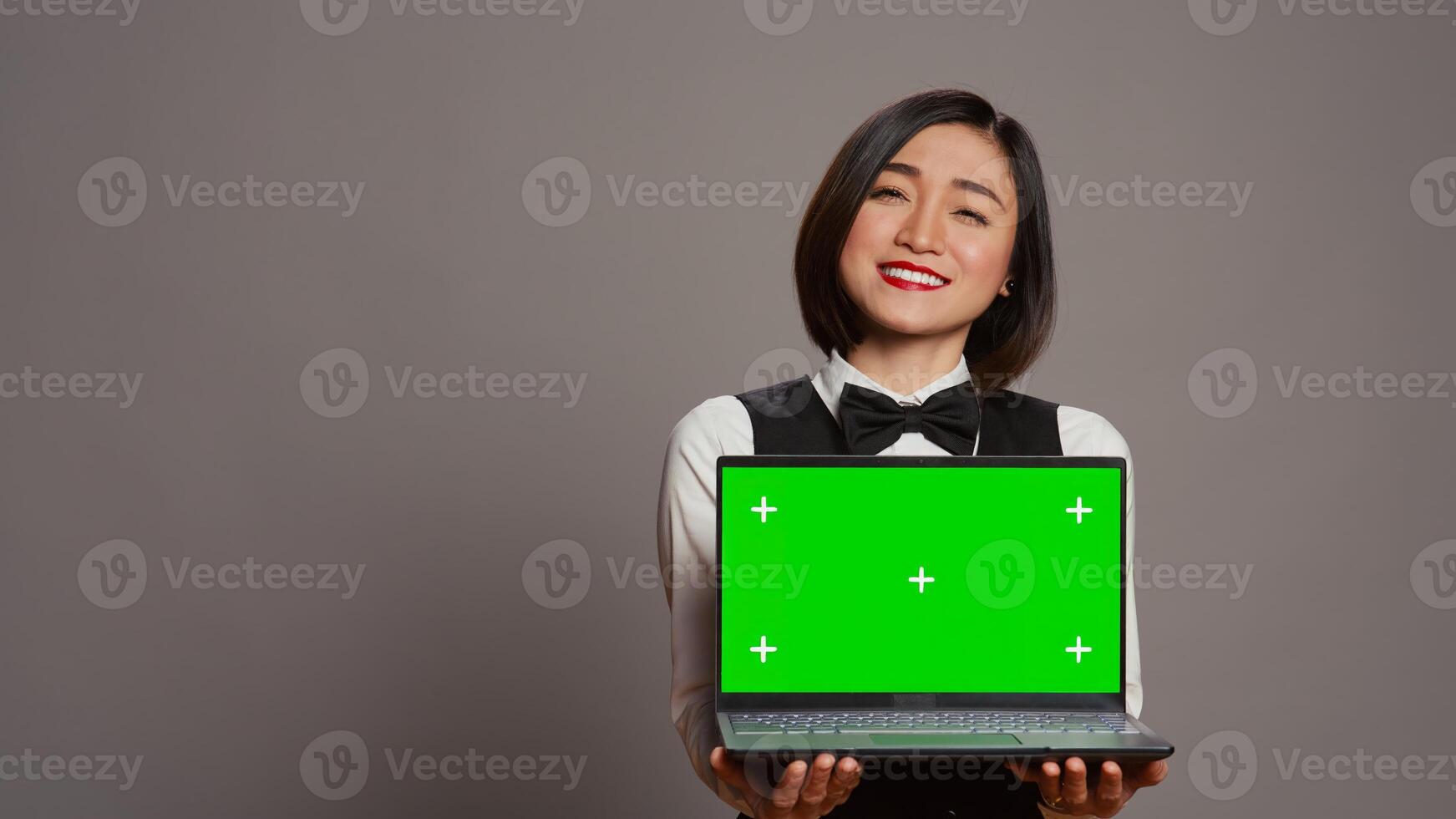 Receptionist showing greenscreen display on personal laptop, presenting isolated copyspace mockup on screen. Asian hotel employee holding pc with chromakey mockup template. Camera B. photo