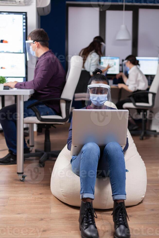 African employee with protection mask against covid-19 pandemic sitting on armchair in middle of office room holding digital tablet. Multiethnic business team working respecting social distance during global pandemic with coronavirus. photo