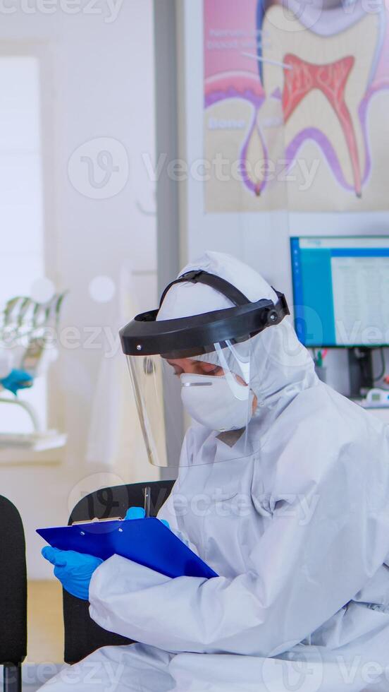 Dentist with face shield talking with elderly woman with toothache before examination during pandemic covid-19 sitting on chairs in waiting area keeping distance. Concept of new normal dentist visit photo