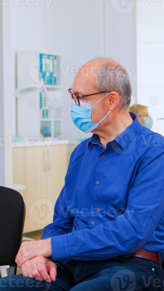 Patients with protection mask talking sitting on chairs keeping social distance in stomatological clinic, waiting for doctor during coronavirus. Concept of new normal dentist visit in covid19 outbreak photo
