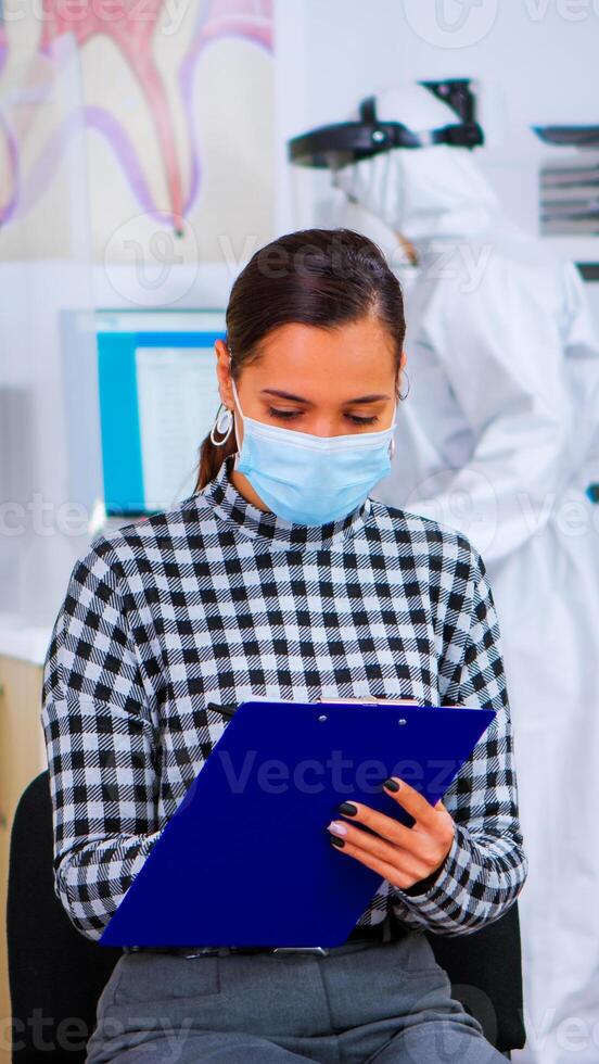 Patient with face protection mask writing on registration form in stomatomoly clinic, sitting in reception respecting social distance. Assistant in coverall measuring patient temperature photo