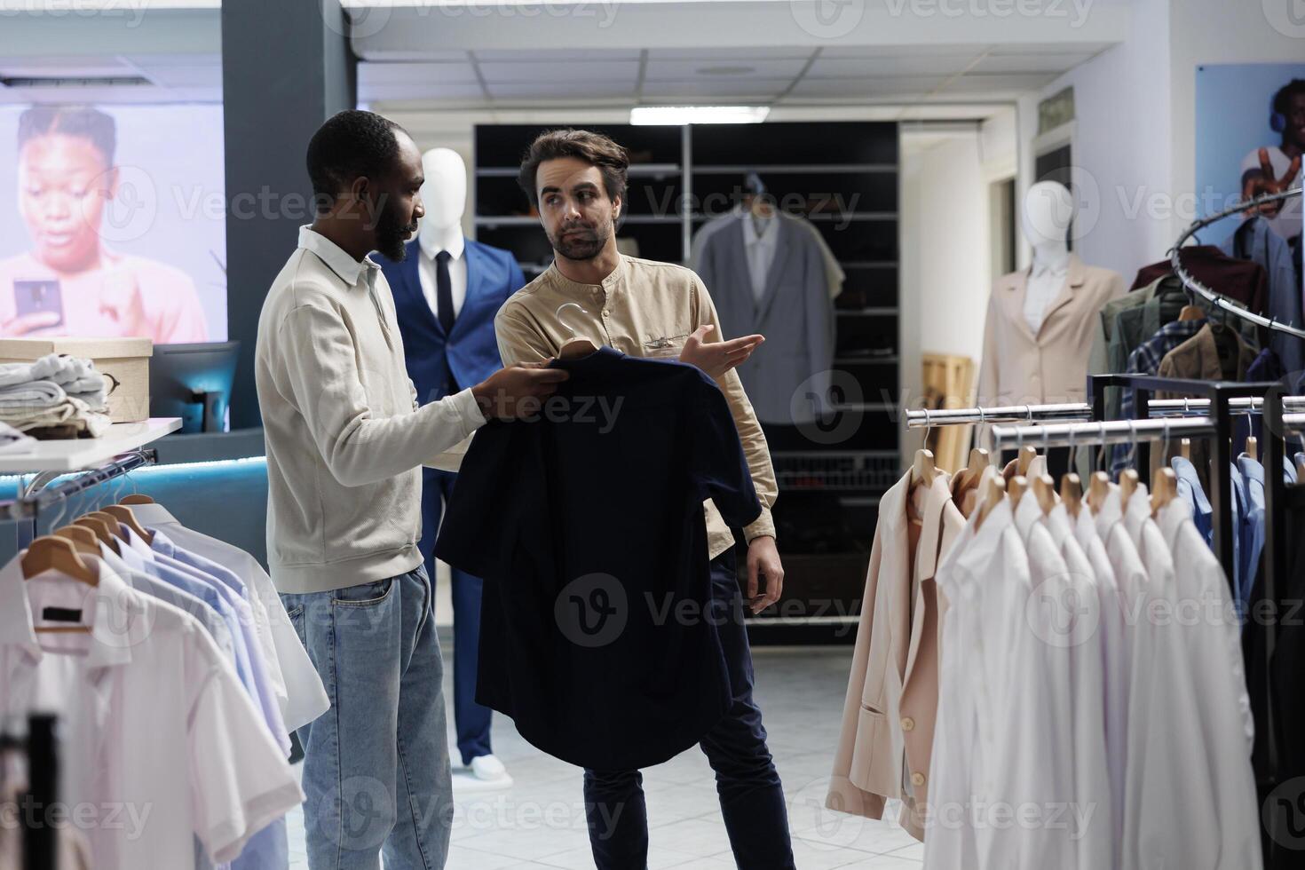 African american man holding shirt on hanger and asking shopping center worker about size options. Boutique worker pointing at new collection apparel rack, helping customer to choose outfit photo