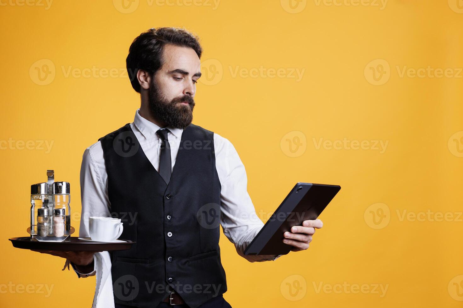 Young employee review list of clients on tablet, holding platter with cutlery in restaurant industry. Skilled waiter having meal tab on device, giving bill to customers on studio camera. photo