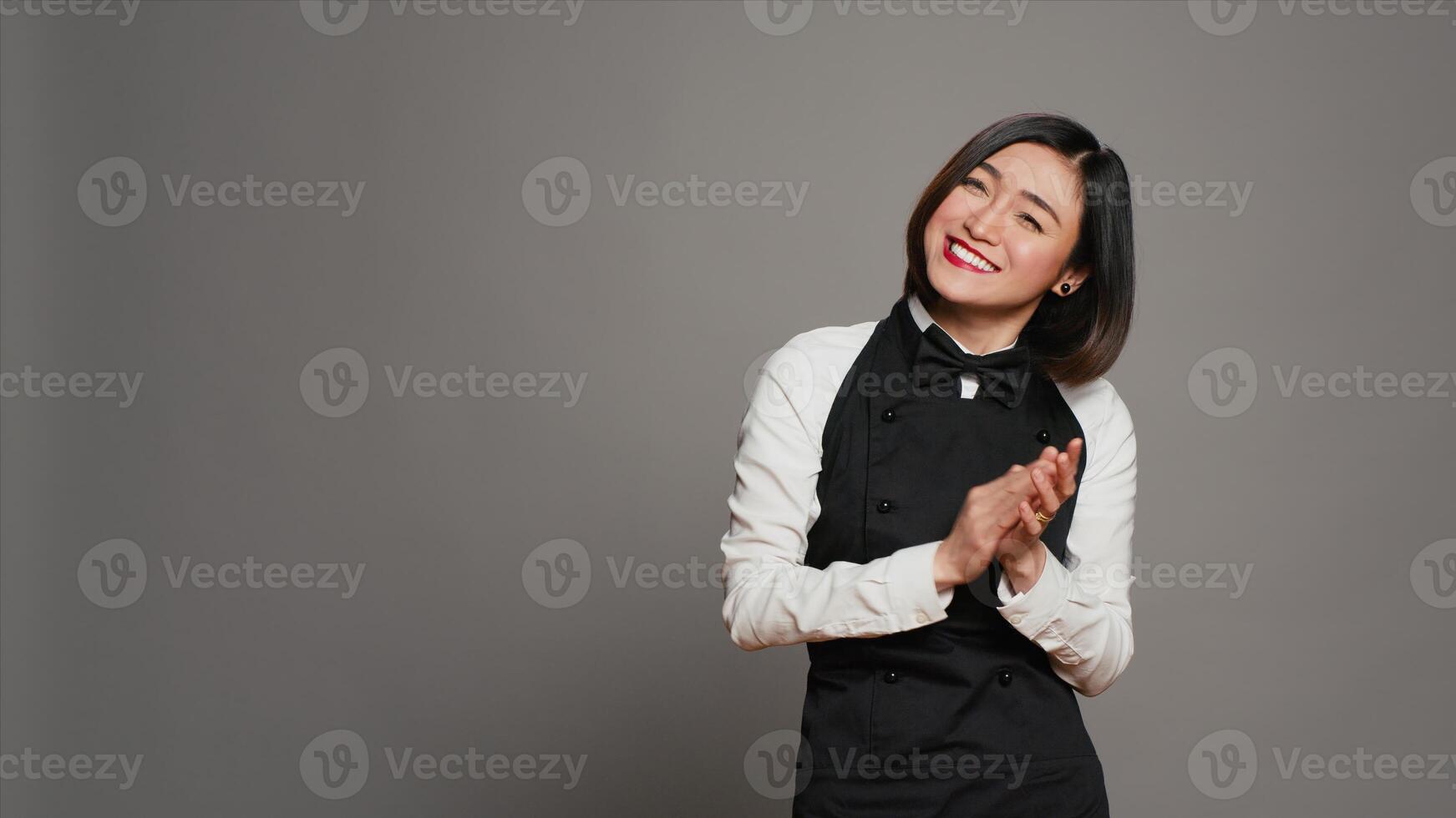 Restaurant hostess clapping hands and celebrating for someone, cheering and saying congratulations in studio. Asian waitress with uniform and apron applauding a person, acclaim. Camera A. photo