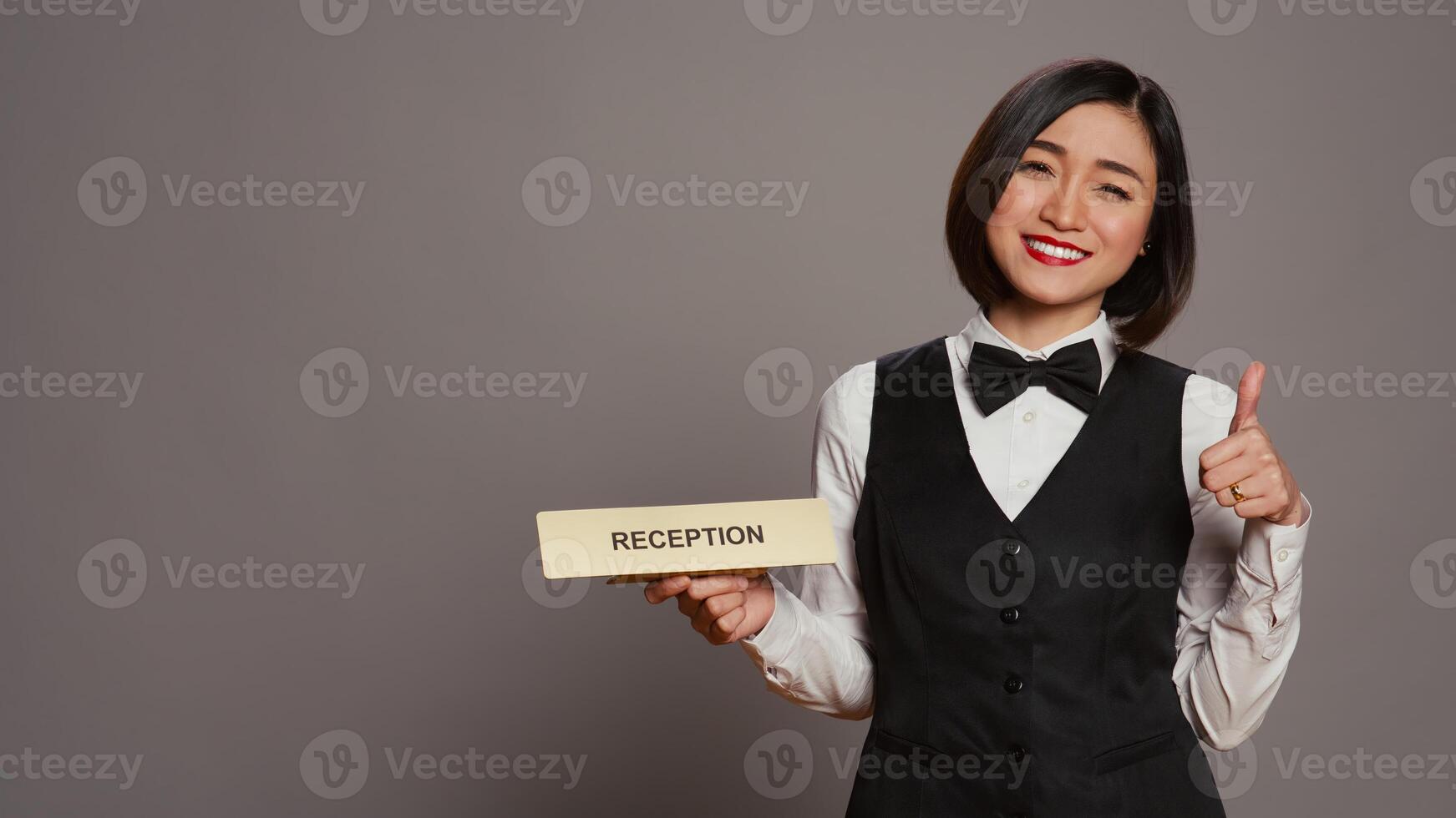 Hotel concierge holding sign to indicate direction for reception desk, presenting indicator to help guests and offer assistance in studio. Employee with uniform and bow uses wall pointer. Camera B. photo