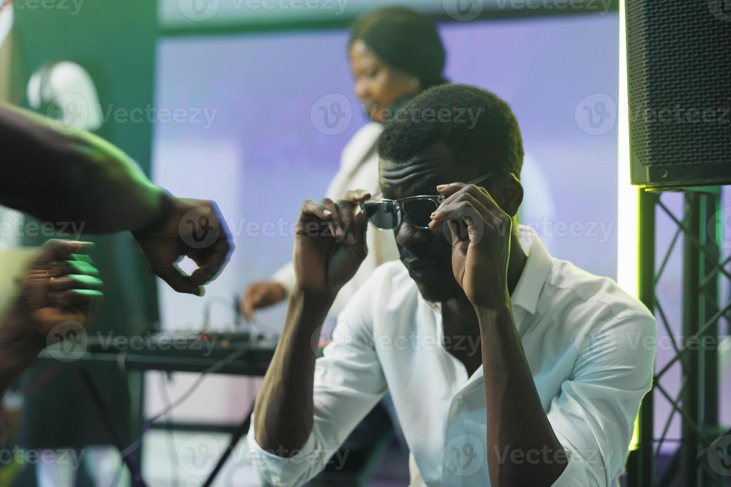 Young african american man wearing sunglasses while sitting in nightclub. Clubber putting on glasses while attending disco party with live electronic music and enjoying nightlife activity photo