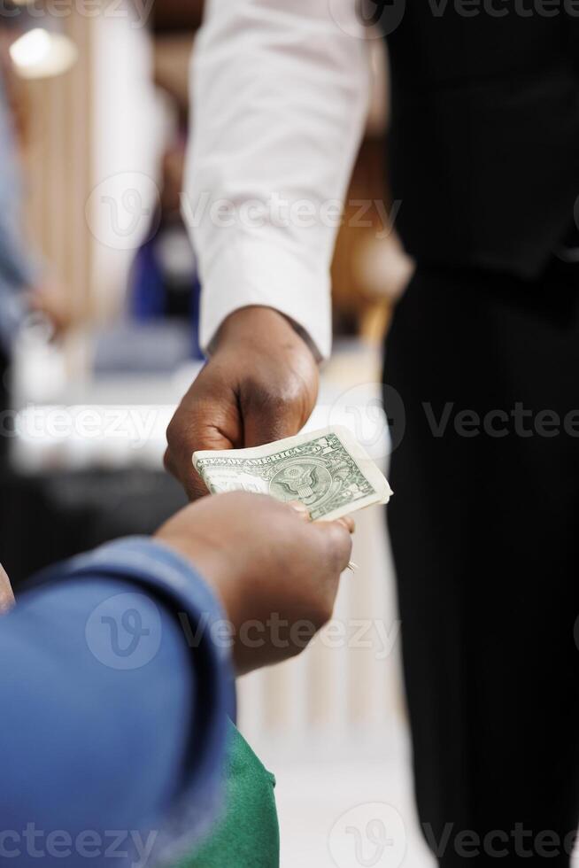 cerca arriba de masculino africano americano camarero recepción propina desde generoso hotel invitado. turista dando dinero efectivo a restaurante obrero, haciendo orden, pago factura. propina en hospitalidad industria foto