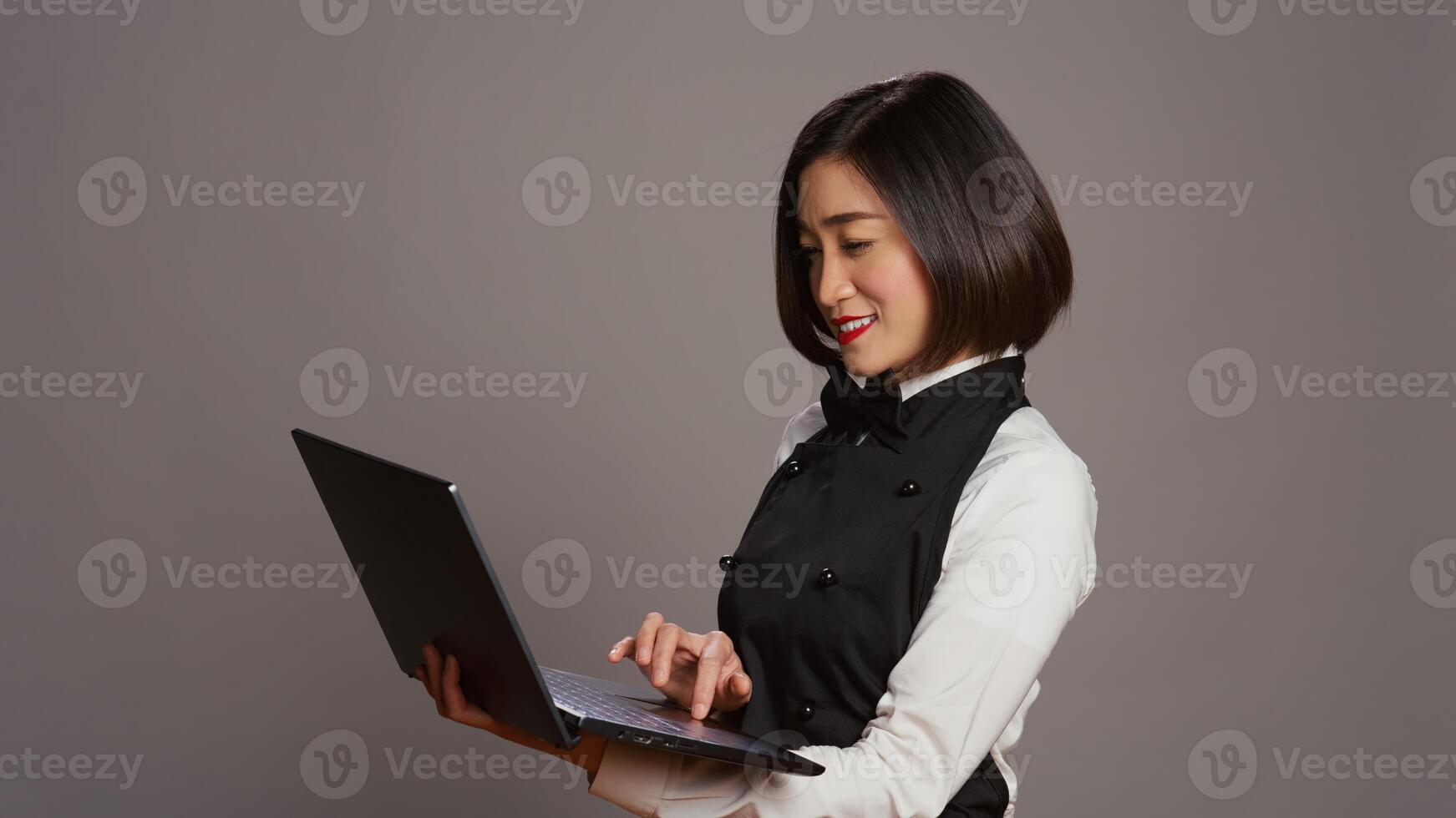 Asian waitress checking all table reservations online on laptop, creating a list of customers and menus at restaurant. Catering employee with apron using pc to offer great service. Camera B. photo