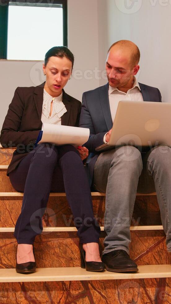 negocio personas mecanografía en ordenador portátil leyendo documentos desde portapapeles analizando informes sentado en escalera en Finanzas corporativo compañía. grupo de profesional gente de negocios caminando en financiero lugar de trabajo. foto