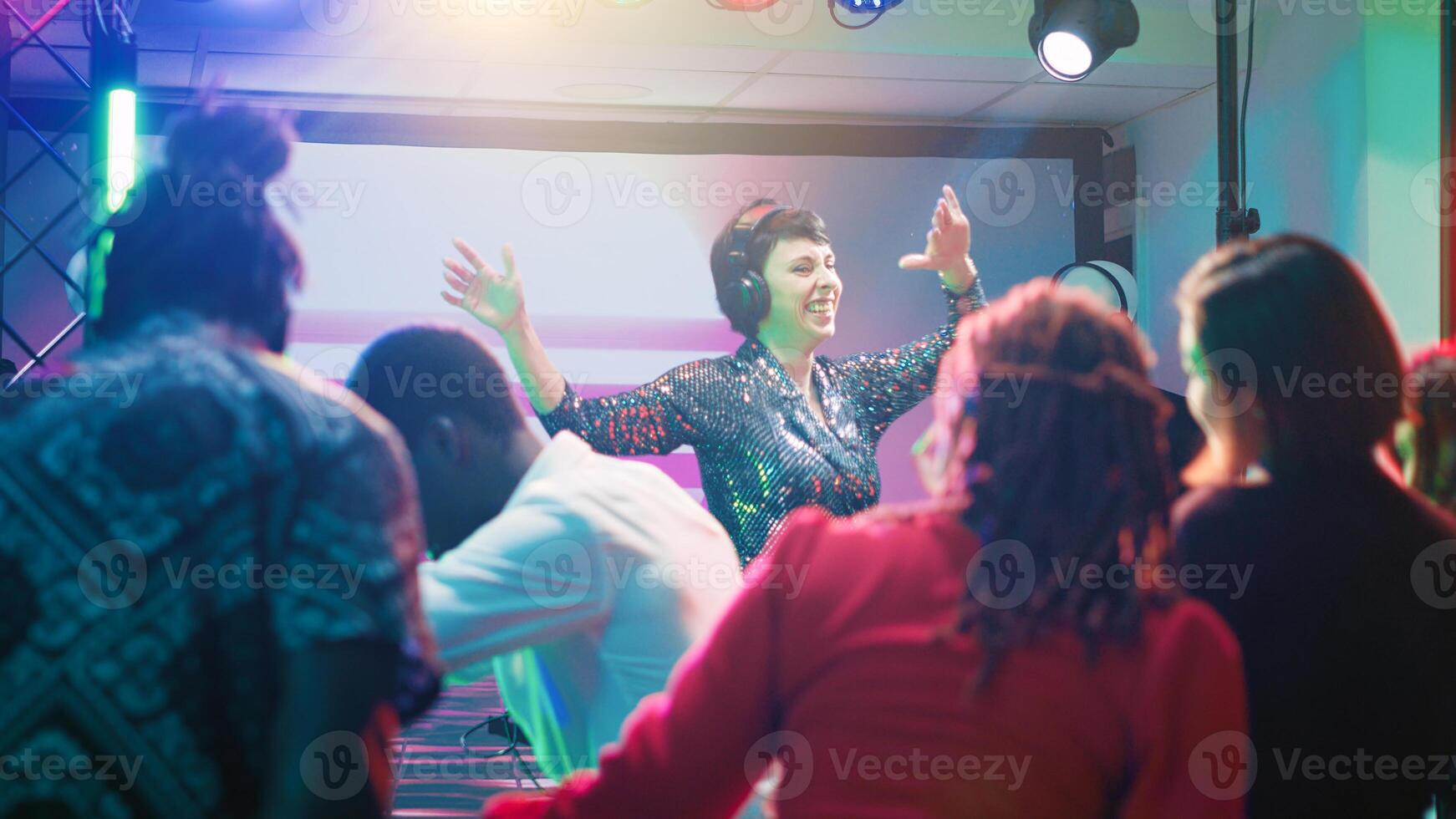 Young DJ mixing funky music at party, feeling cheerful with dance moves and electronic sounds at station on stage. Group of friends partying and dancing together at club, nightlife. photo
