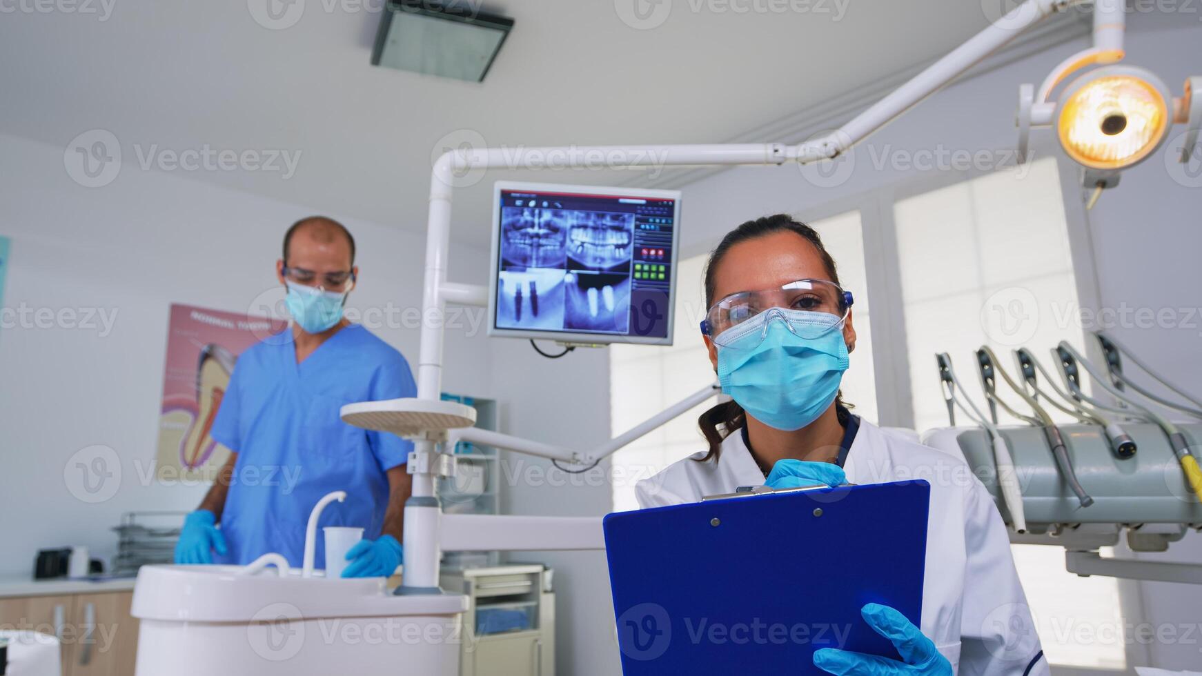 odontología médico interrogando paciente y tomando notas en portapapeles antes de cirugía. médico y enfermero trabajando en moderno ortodoncia oficina, escritura y examinando persona vistiendo proteccion máscara foto