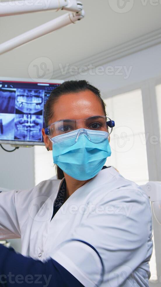 POV of patient in a dental clinic sitting on surgery chair checking affected mass. Dentistry team working in orthodontic office, lighting the lamp and examining person, close-up face in medical mask. photo