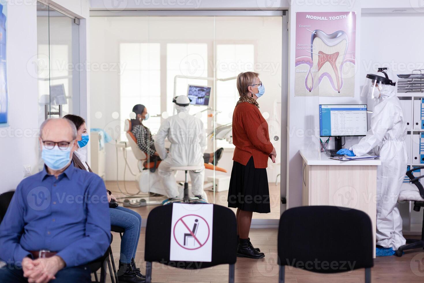 Senior patient with face msak discussing with dentist assistant dressed ppe suit keeping social distancing in waiting area. Dentistiry staff in protection suit during global pandemic with covid-19. photo