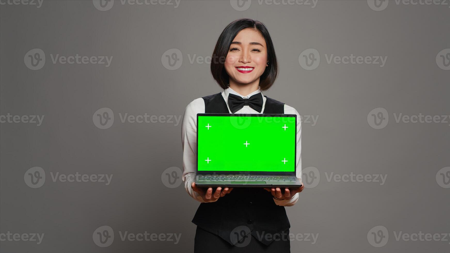 Asian hotel concierge holding pc with greenscreen on camera, presenting isolated mockup template on screen. Receptionist showing laptop with blank chromakey layout on display. Camera A. photo