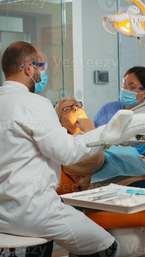 Dentist treating teeth to senior woman patient in clinic lying on chair with open mouth. Doctor and nurse working together in modern stomatological office wearing protection mask photo