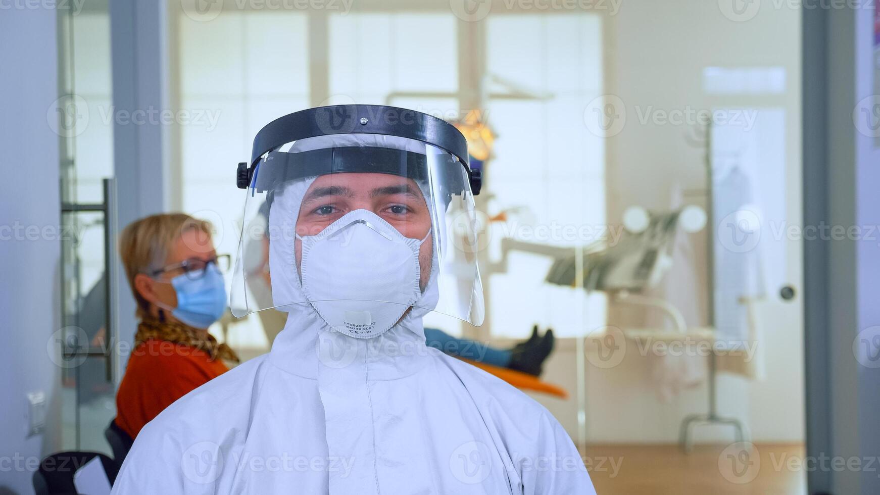 Portrait of exhausted doctor in dental office looking on camera wearing ppe suit and face shield sitting on chair in waiting room clinic. Concept of new normal dentist visit in coronavirus outbreak. photo