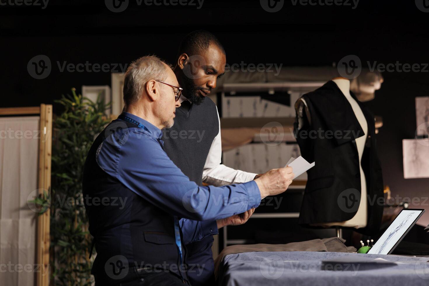 Master menswear collection creator and apprentice working on sartorial suit adjustments. Tailors modifying fashion sketches, trying different clothing combinations on mannequin in atelier photo