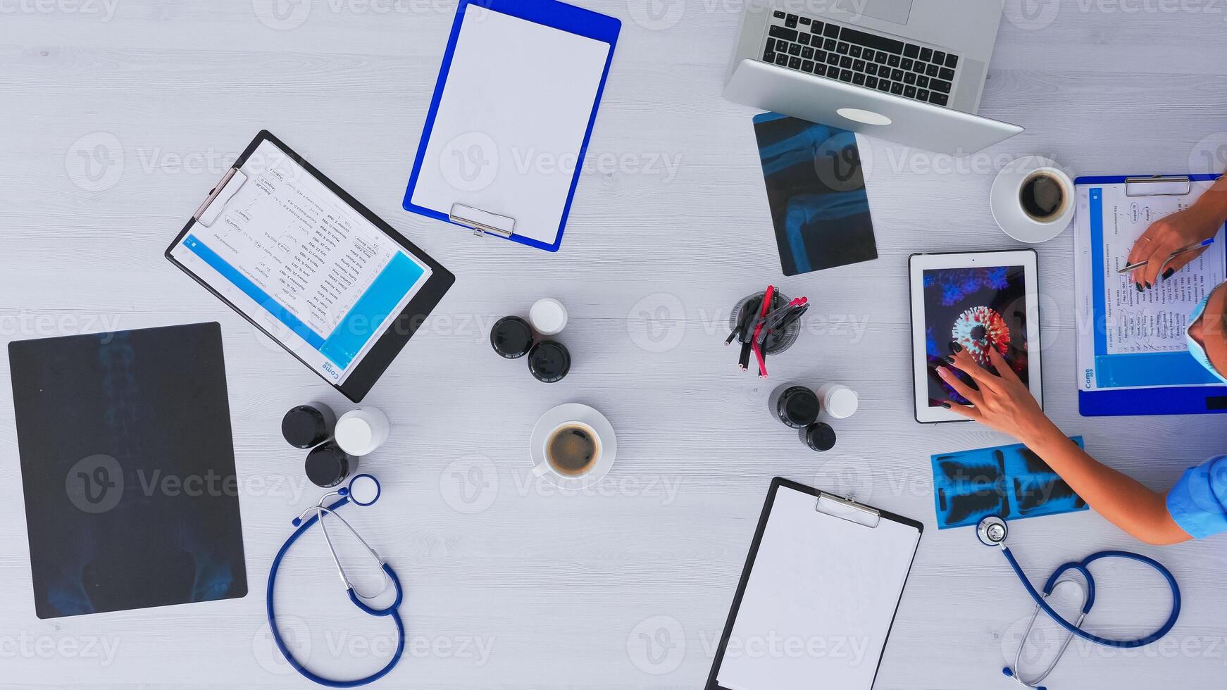 Top view of doctor researching about virus using tablet, taking notes sitting on desk in medical clinic with x-ray and digital technology. Assistant writing on clipboard, copy space, flat lay concept. photo