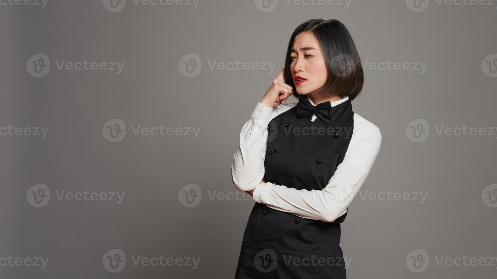 Asian waitress thinking about new menu ideas for restaurant, acting pensive in front of camera. Gourmet employee being creative with food plates, thoughtful server with apron and bow. Camera A. photo