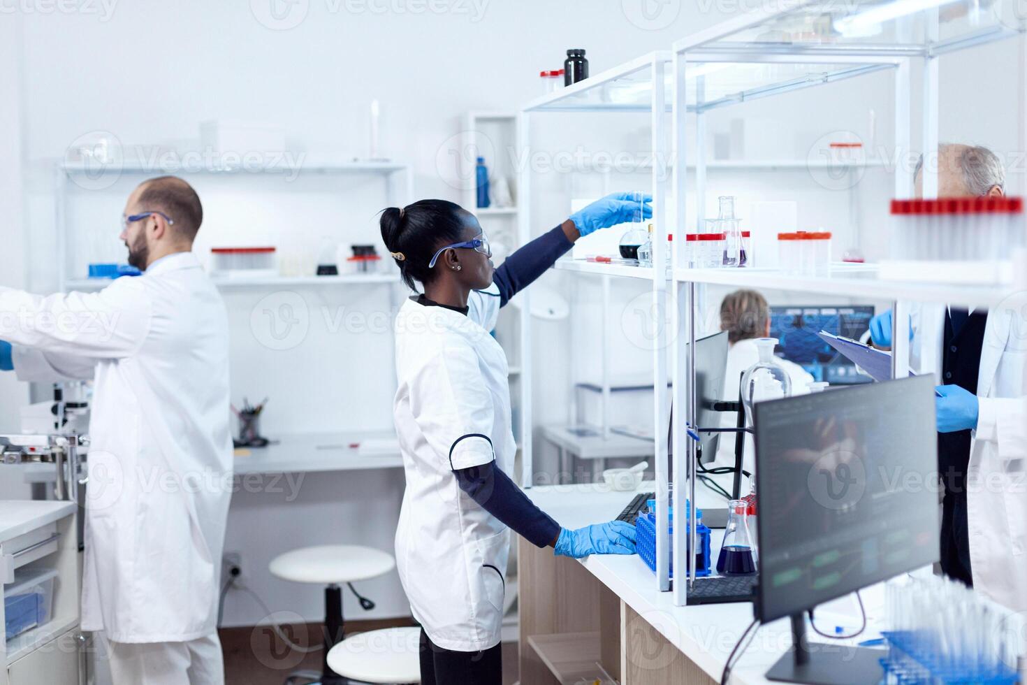 africano en biotecnología laboratorio alcanzando para vaso matraz desde estante. multiétnico equipo de investigadores trabajando en microbiología laboratorio pruebas solución para médico objetivo. foto