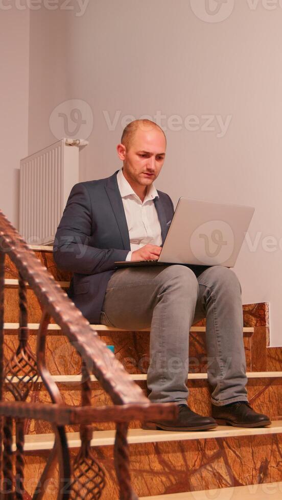 Stressed overworked business man smoking sitting on stairs working on difficult corporate deadline. Entrepreneur doing overtime at job on staircase while colleagues leaving office building. photo