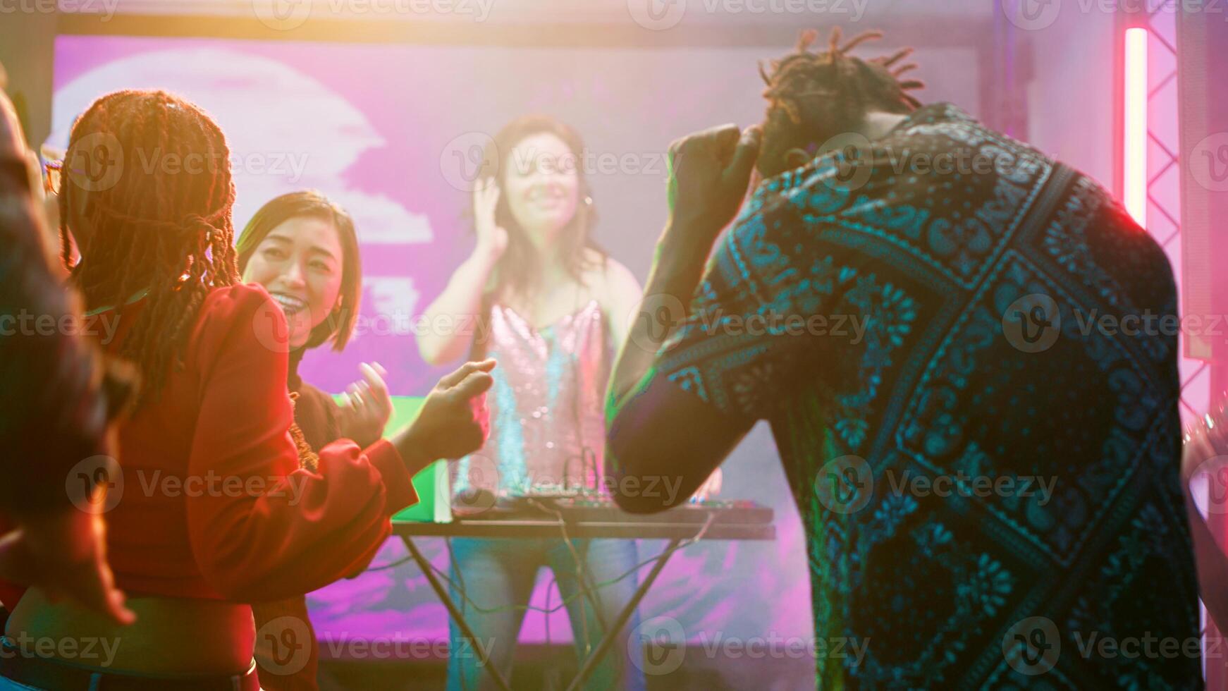 Diverse group of friends partying on dance floor, woman DJ mixing sounds on stage at discotheque. Cheerful people dancing on funky music at nightclub, having fun with bright lights. photo