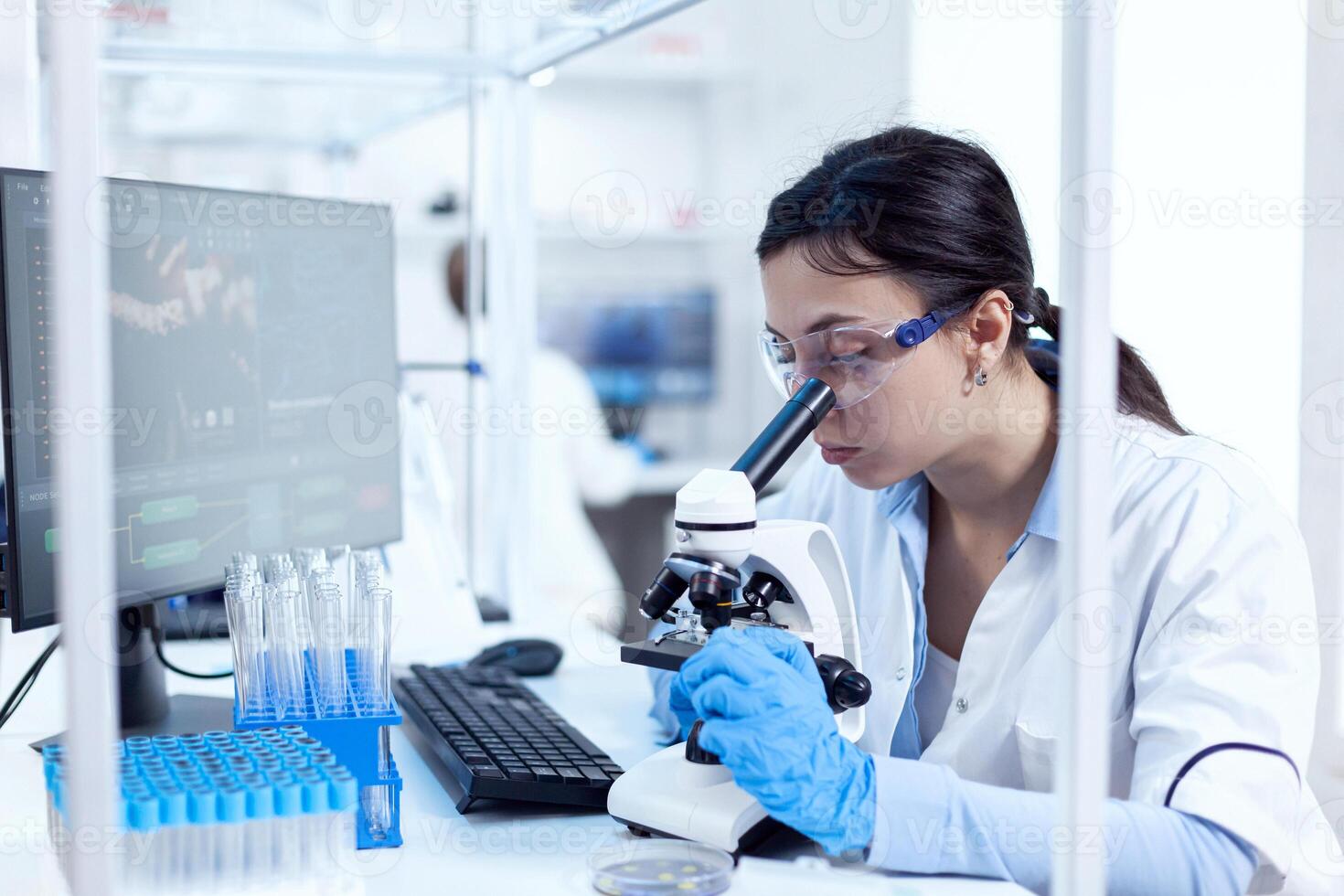 Microbiologist in research laboratory doing scientific experiments looking through microscop. Chemist wearing lab coat using modern technology during scientific experiment in sterile environment. photo