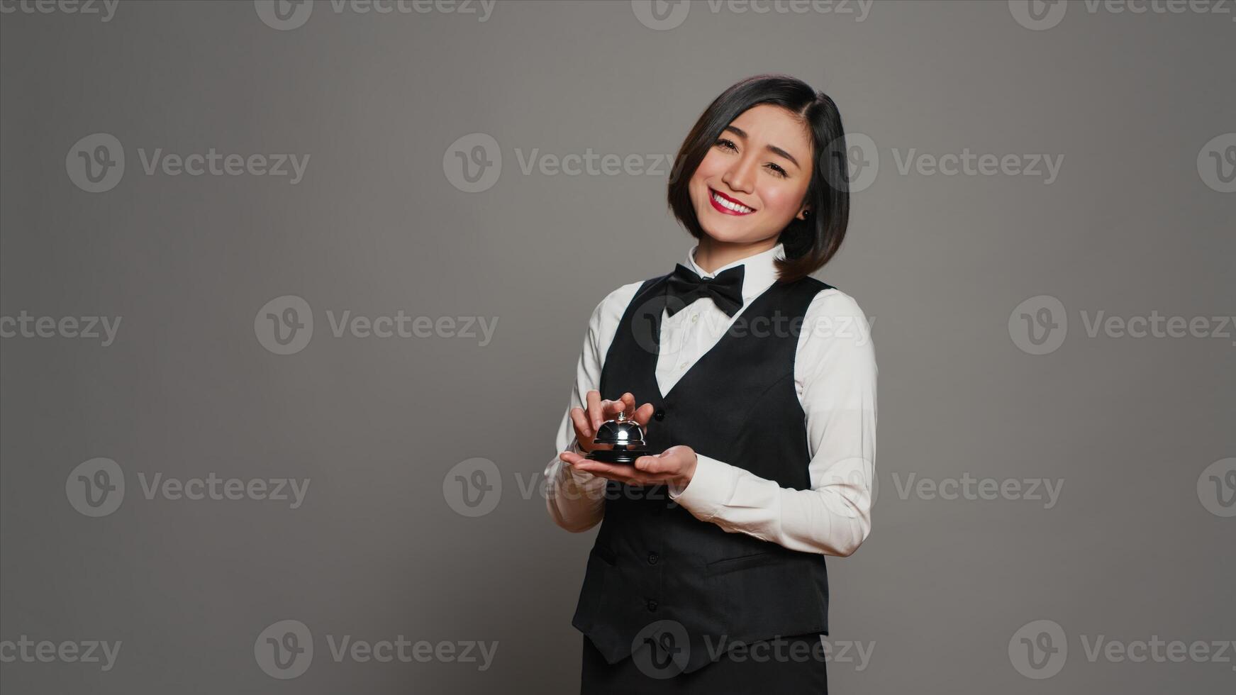 Woman hotel employee ringing front desk service bell in studio, working as a receptionist and registering guests in rooms. Asian concierge presenting a bell to call for assistance. Camera A. photo