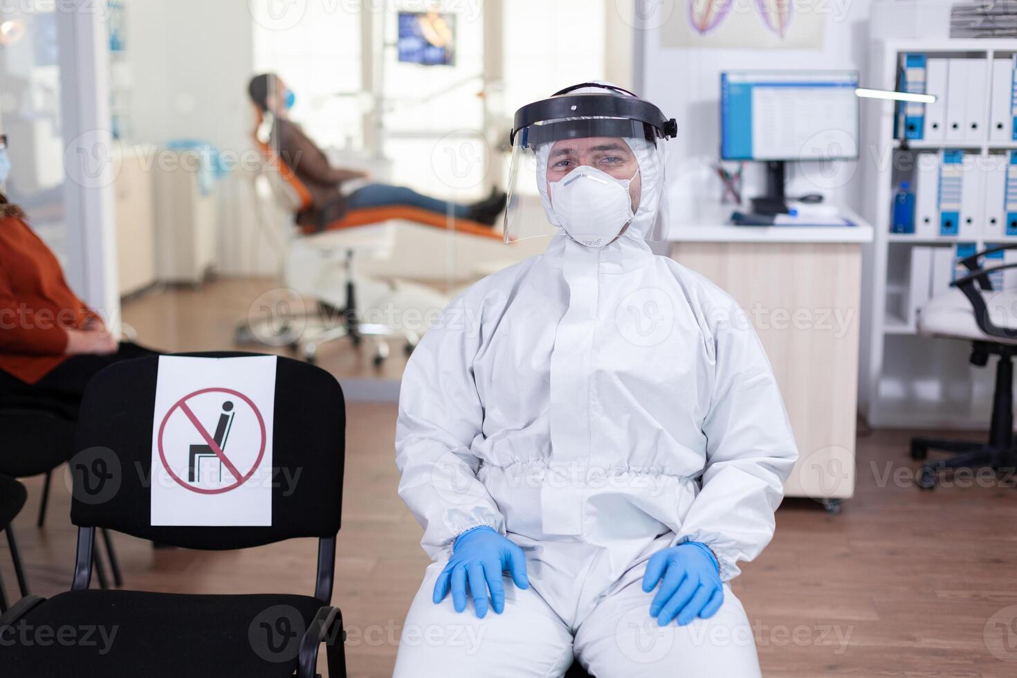 Close up of tired doctor in stomatological office looking on camera wearing overall and face shield sitting on chair in waiting room clinic. Concept of new normal dentist visit in coronavirus outbreak. photo