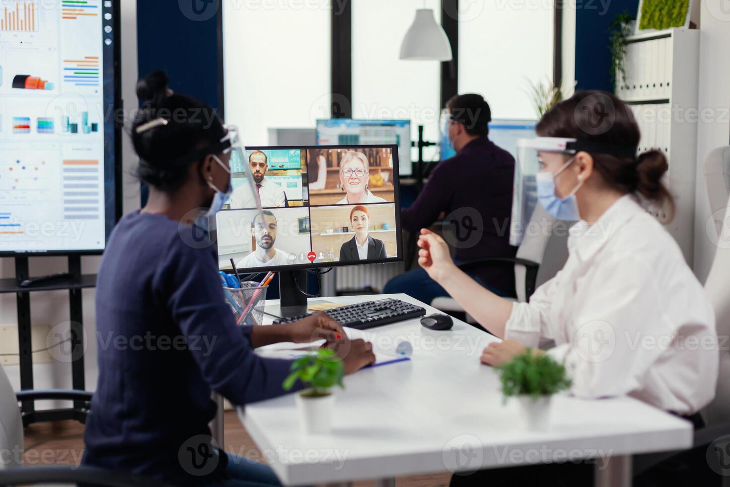 Black woman and colleague listening business people on video call wearing face mask during covid19. New normal business office. Multiethnic team working respecting social distance during global pandemic with coronavirus. photo