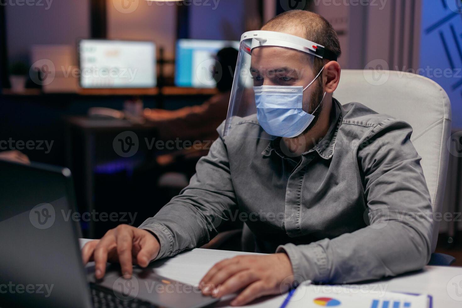 Businessman protect himself with visor against covid-19 late in the office distancing from colleagues. scrolling on laptop. Stressed man in corporation working hard to finish a project wearing face mask as safety precaution due to coronavirus pandemic. photo