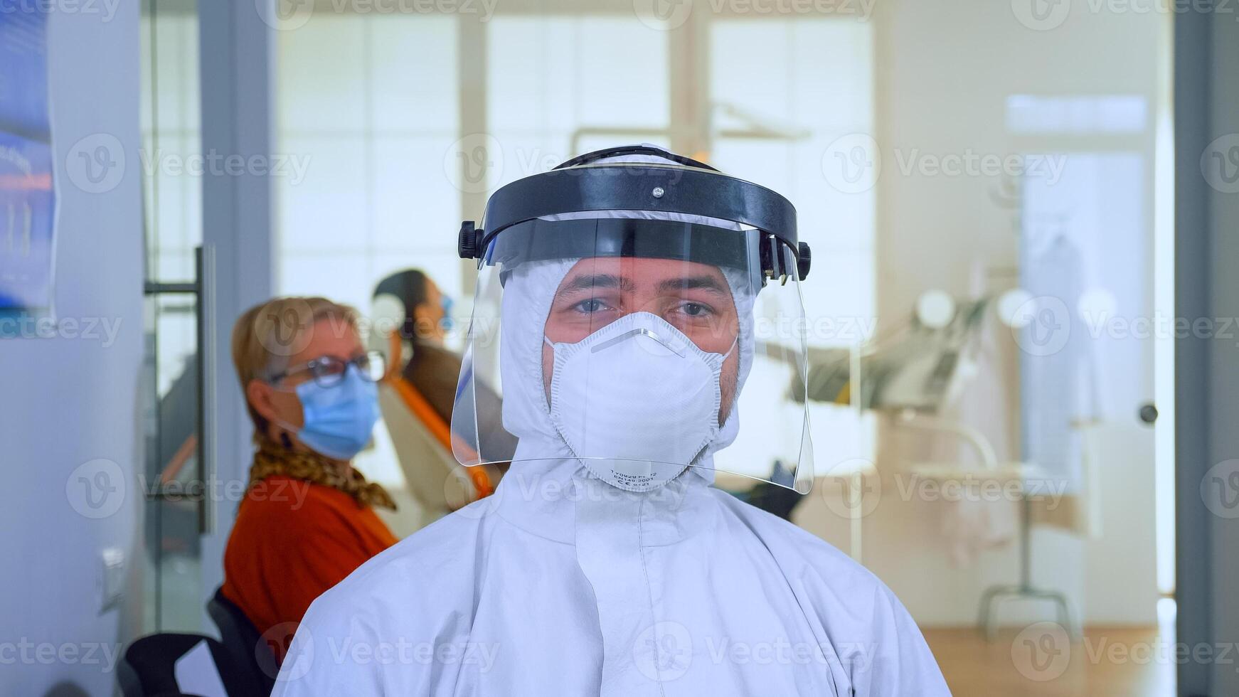 Close up of tired doctor in stomatological office looking on camera wearing overall and face shield sitting on chair in waiting room clinic. Concept of new normal dentist visit in coronavirus outbreak. photo