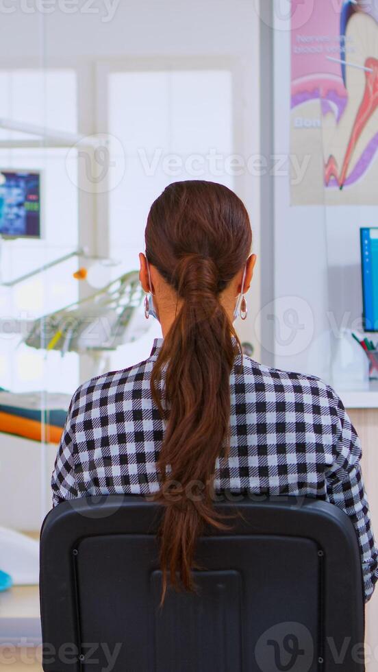Assistant with face mask against covid-19 coming in reception typing on computer while patients sitting on chairs in waiting room talking keeping distance. Concept of new normal dentist visit. photo