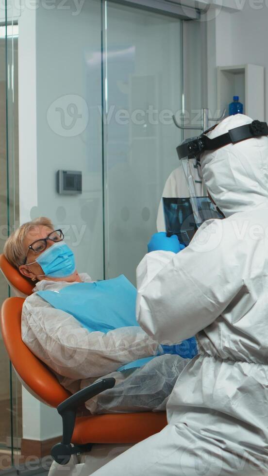 Stomatologist in protective suit asking for patient dental x-ray examining teeth problems during coronavirus pandemic in modern clinic. Medical team wearing coverall, face shield, mask and gloves. photo