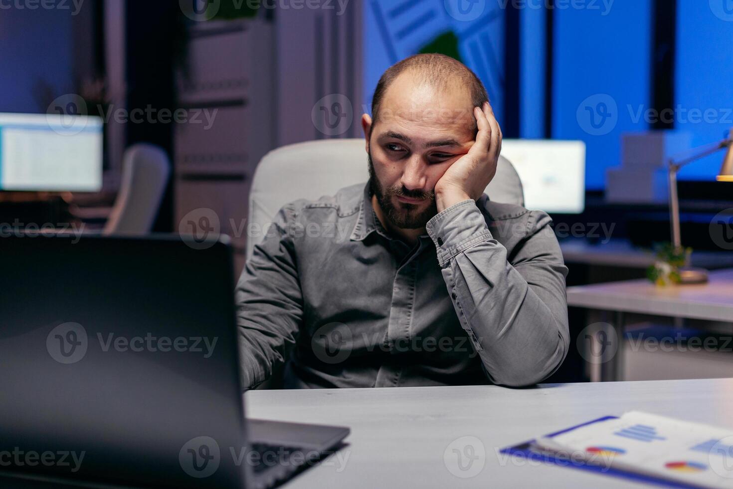 Tired businessman sit at the computerin the evening works on deadline. Workaholic employee falling asleep because of working late at night alone in the office for important company project. photo