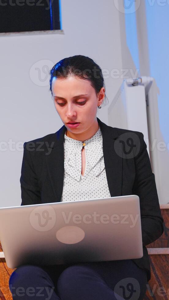 sobrecargado de trabajo estresado mujer de negocios trabajando tarde en noche en negocio fecha límite en ordenador portátil. grave empresario siendo en corporativo trabajo sentado en escalera de negocio edificio con el tiempo. foto