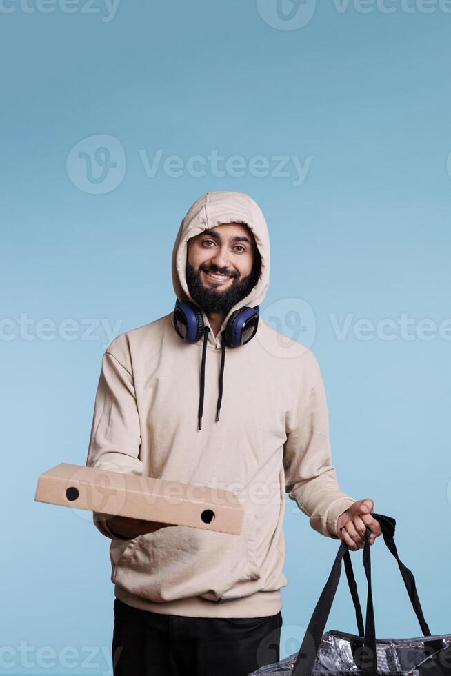 Smiling food delivery service arab courier holding pizza box and backpack portrait. Cheerful young man in hood giving fastfood takeaway order and looking at camera with carefree expression photo