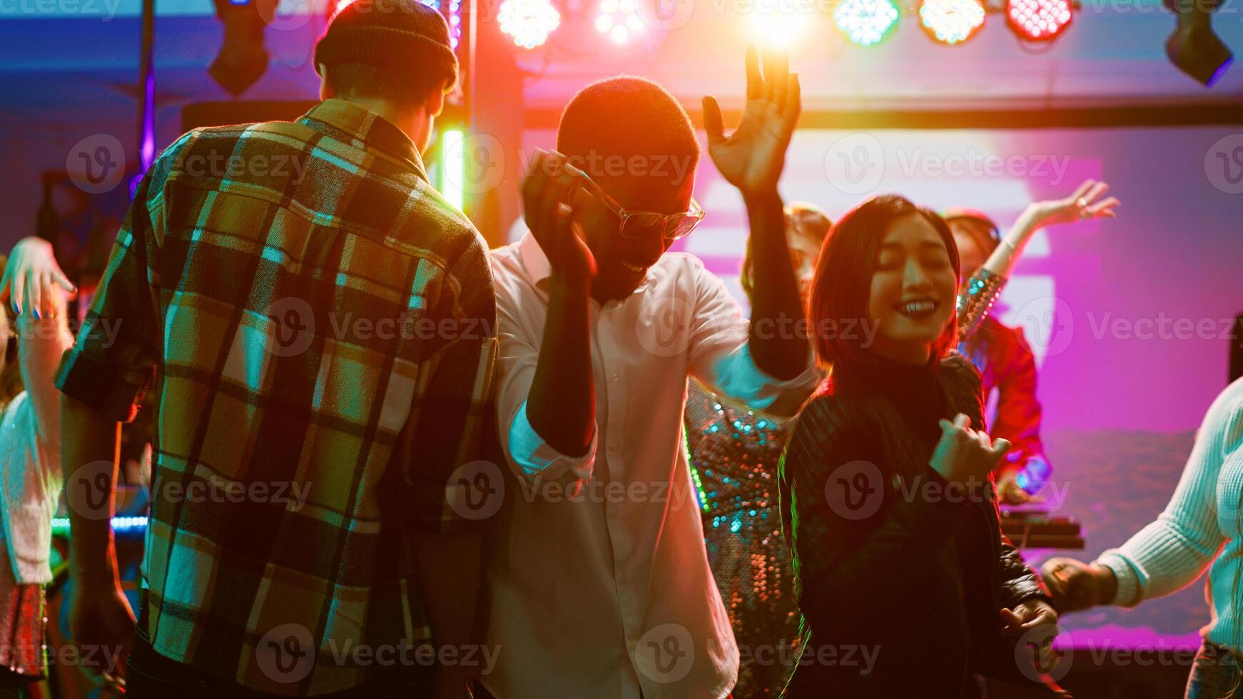 Diverse friends dancing on live music, feeling cheerful on dance floor at nightclub. Young people having fun with performance at discotheque, doing funky dance moves under stage lights. Tripod shot. photo