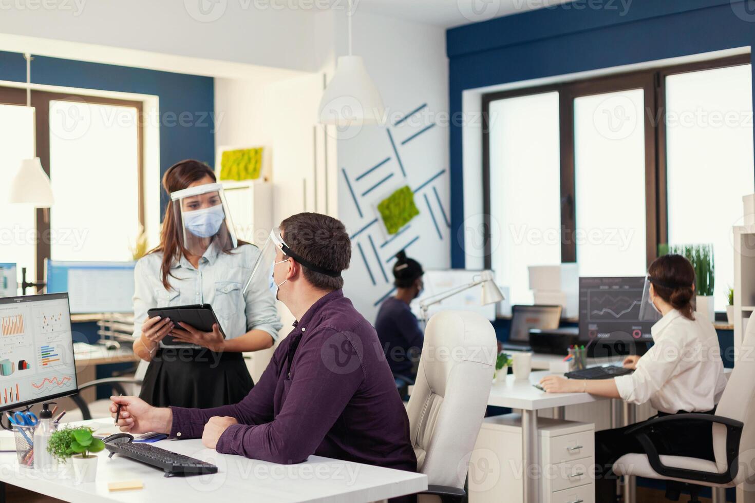 Business team helping each other to finish a deadline at workplace wearing face mask during covid19. Multiethnic team in new normal financial office in corporate company typing on computer, taking notes on tablet. photo