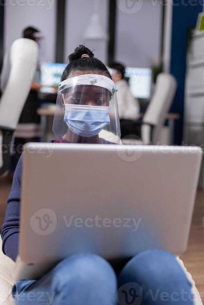 African businesswoman working on laptop wearing face mask against covid19. Multiethnic business team working respecting social distance during global pandemic with coronavirus. photo